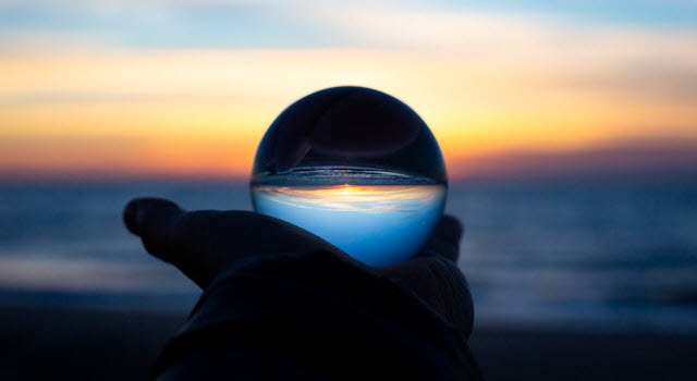 A silhouetted hand holds a clear glass orb at arm's length, with a vivid sunset over the ocean in the background. The glass sphere inverts and distorts the view, showing an upside-down image of the glowing horizon and sky within its surface.